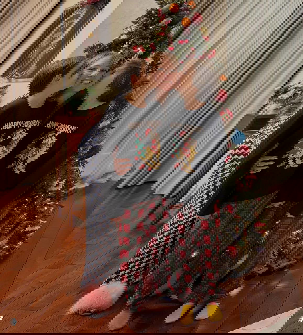 Мама с двумя сыновьями у небольшой украшенной ёлки: тёплые объятия, одинаковые праздничные свитеры и деревянный пол в интерьере.