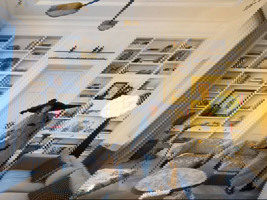 Широкий план гостиной с высокими белыми книжными полками, мягкими креслами с бахромой, низкими мраморными столами и необычным торшером в углу