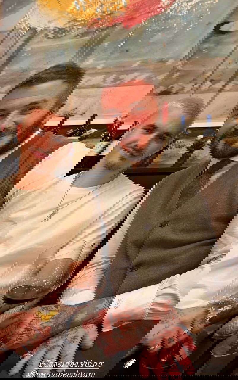 Два человека сидят за столом в ресторане с бокалами вина; интерьер в тёплых тонах и акцент на руках с татуировками и украшениях.