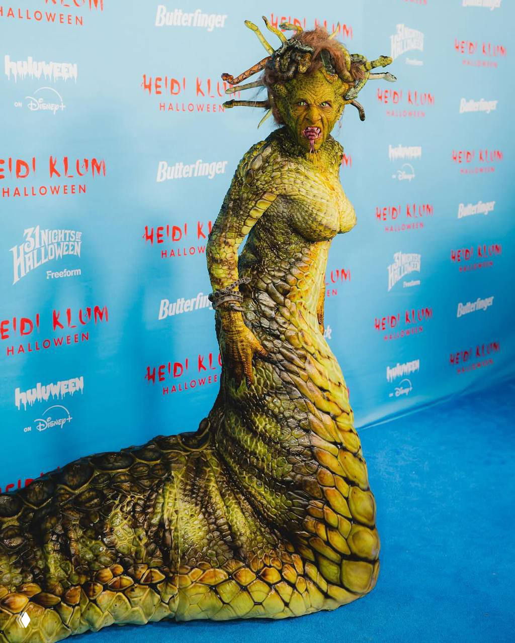 Полный рост Хайди Клум в образе Медузы Горгоны: длинный 2,5-метровый хвост костюма, чешуйчатая текстура и синяя фотозона на фоне.