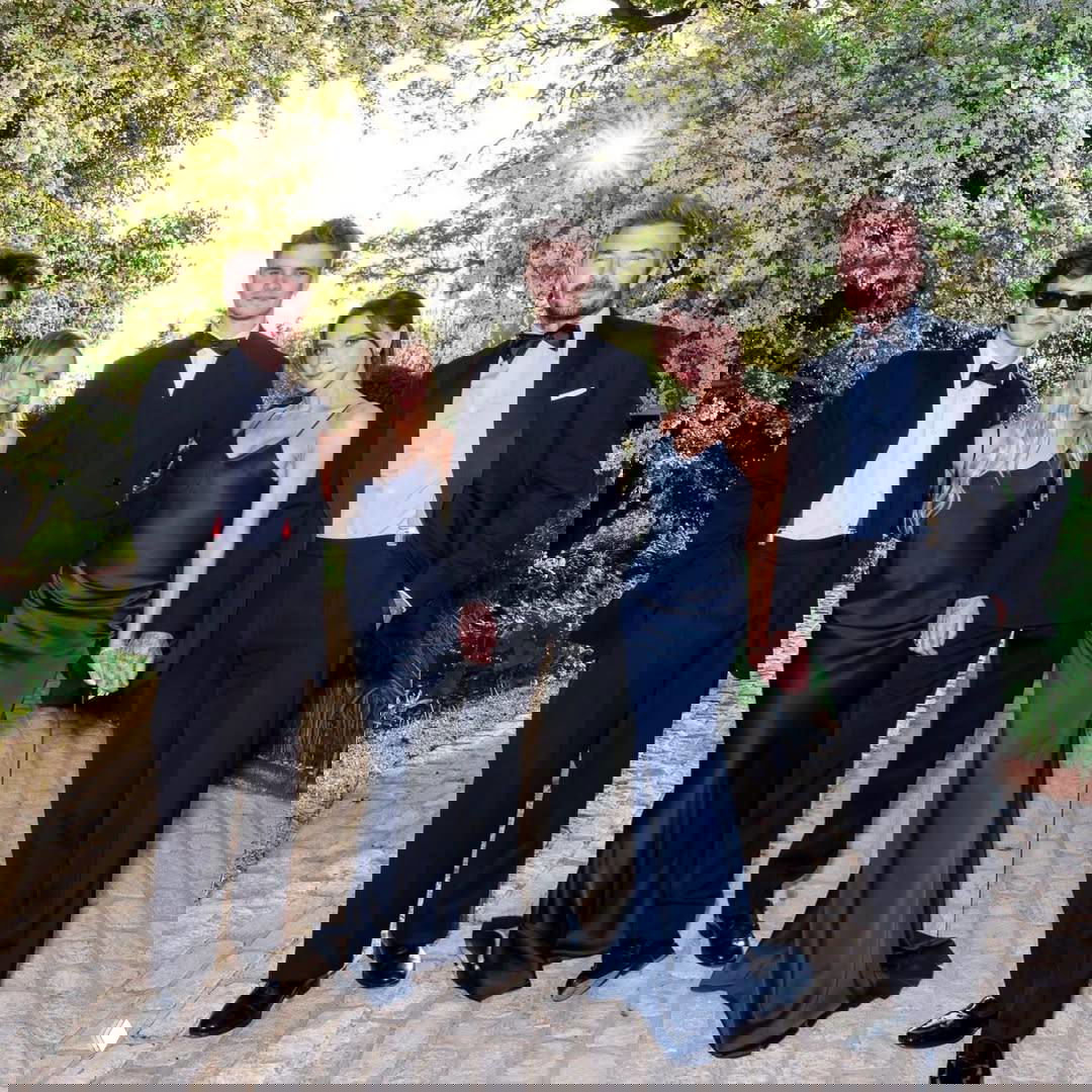 Группа людей в смокингах и вечерних платьях позируют вместе на дорожке посреди цветущих деревьев при солнечном свете.