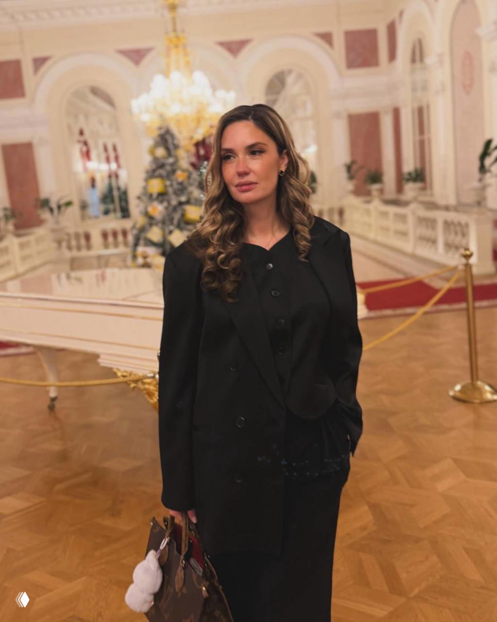 Женщина в чёрном костюме на фоне рождественской ёлки и роскошного фойе Большого театра, классическая праздничная обстановка.