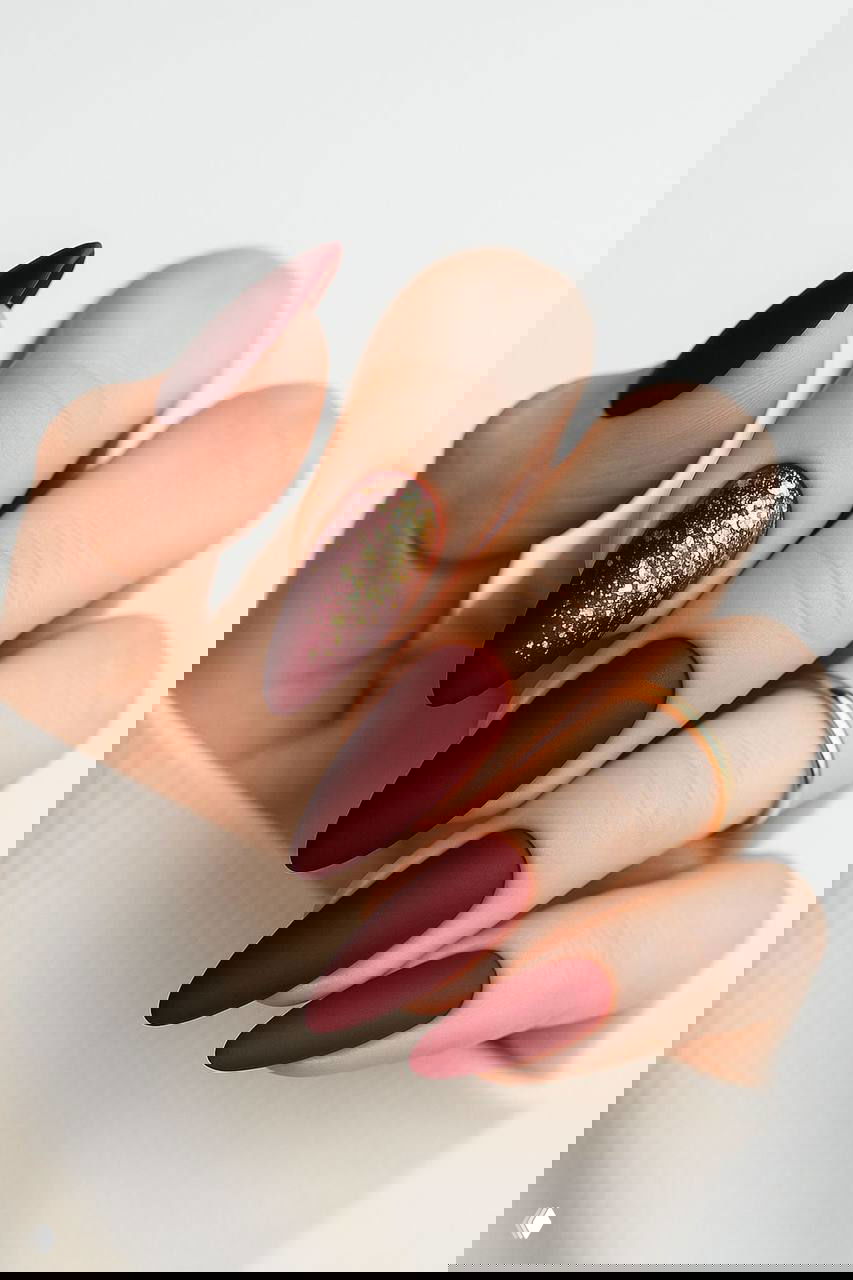 Матовый бордово-красный удлинённый маникюр с золотыми блёстками на одном ногте и элегантными кольцами на пальцах — трендовый новогодний вариант.