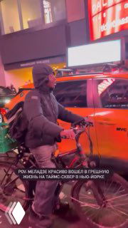 Мужчина на велосипеде рядом с жёлтым такси в ярко освещённой части Таймс-сквер, кадр из видео с концерта в Нью‑Йорке, ночная городская сцена