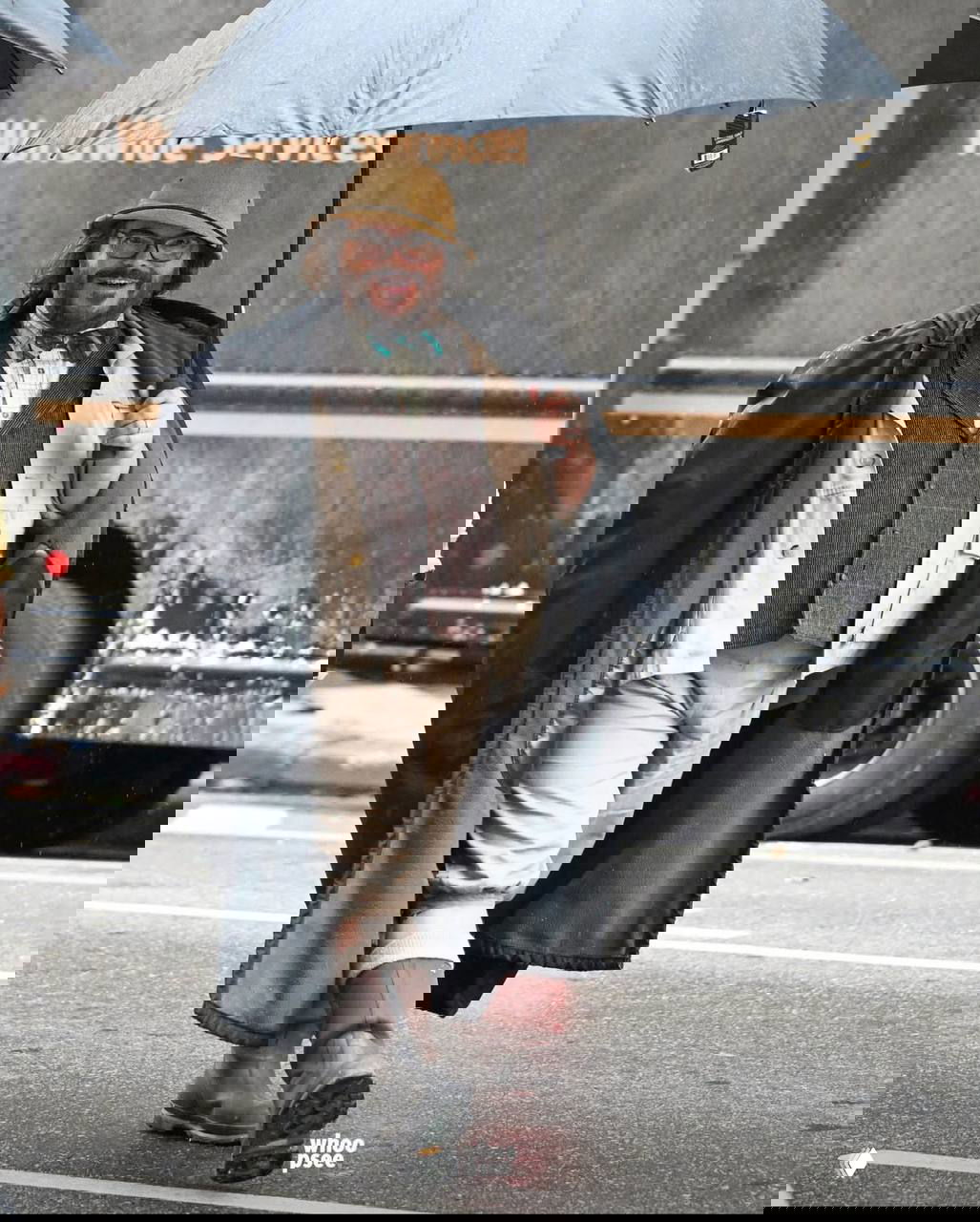 Актёр в шляпе с зонтом и винтажным костюмом идёт по мокрой улице рядом с грузовиком — кадр со съёмок фильма с праздничными декорациями.