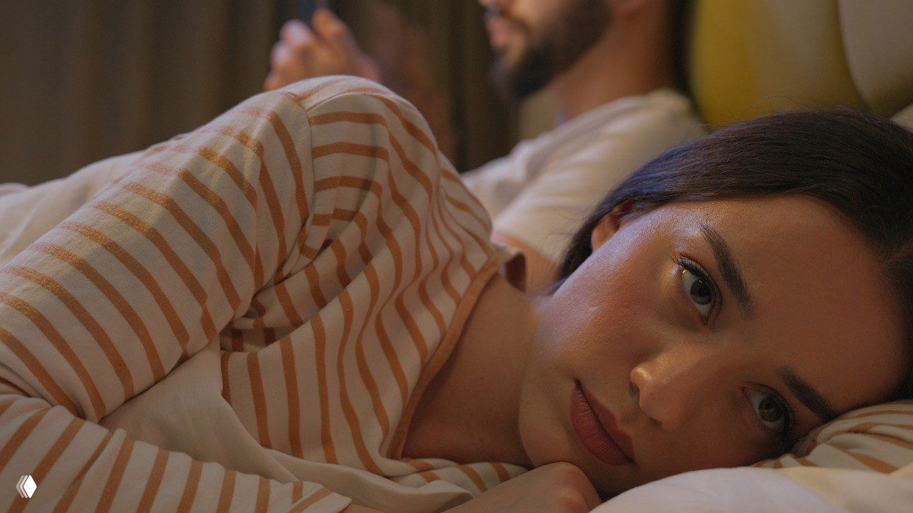 Женщина лежит на кровати в полосатой пижаме, грустно смотрит в сторону; на фоне размыто виден мужчина — символ напряжения и отдаления в паре.