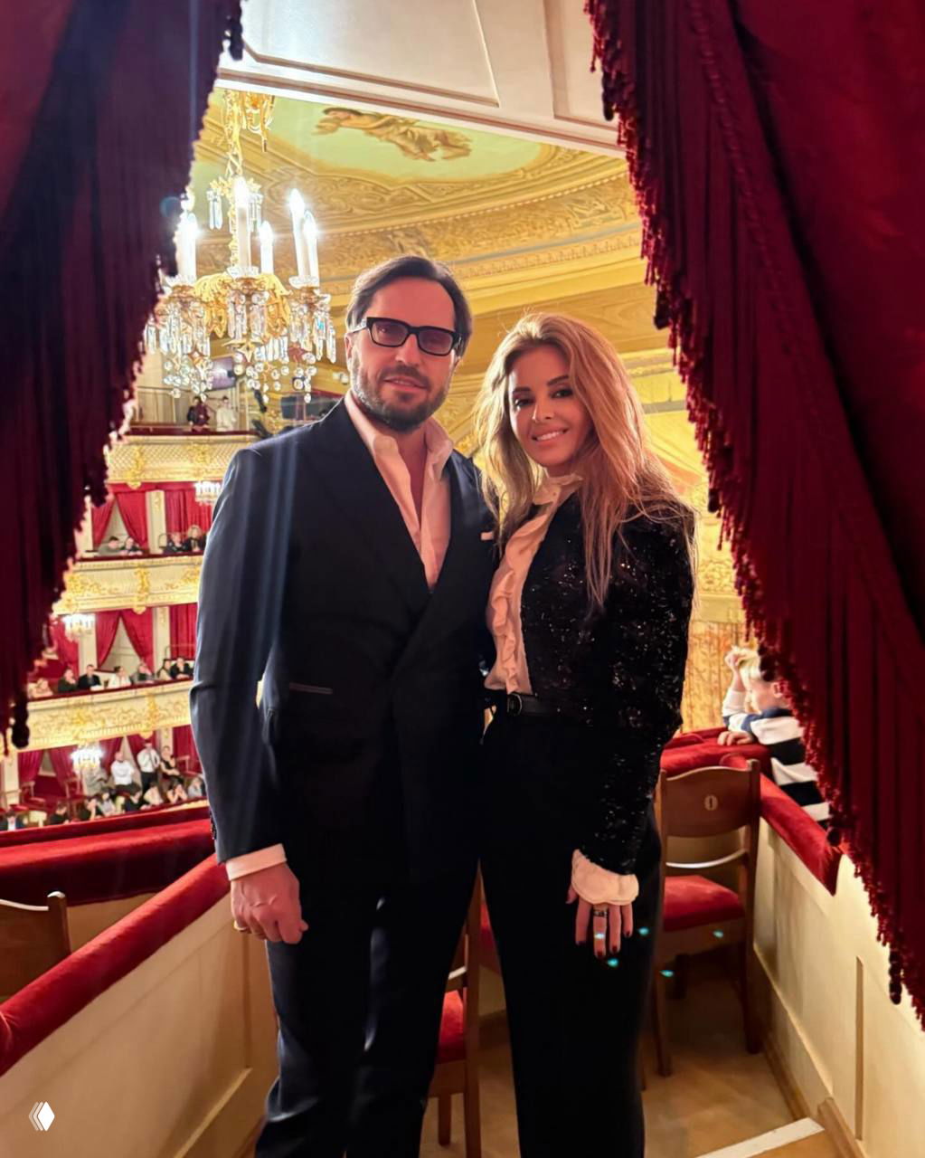 Мужчина в смокинге и женщина в чёрном блейзере позируют в театральной ложе на фоне красных кресел и орнамента Большого театра.