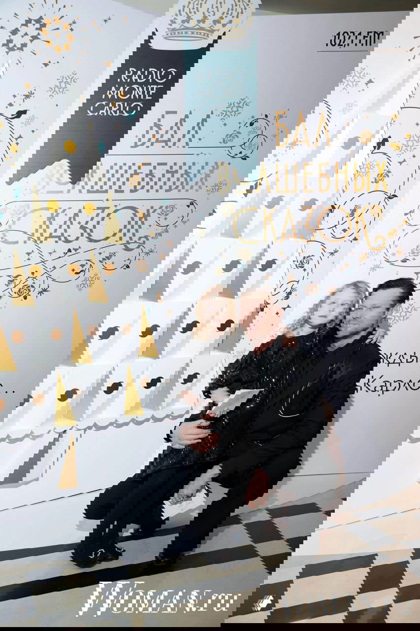 Взрослый мужчина присёл рядом с маленькой девочкой у декоративной ёлки и брендволла Radio Monte Carlo на семейном фоторепортаже бала.