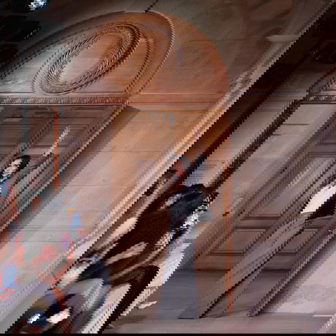 Женщина в брючном костюме выходит на подиум или сцену, вокруг сидит публика, некоторые фотографируют на телефоны.