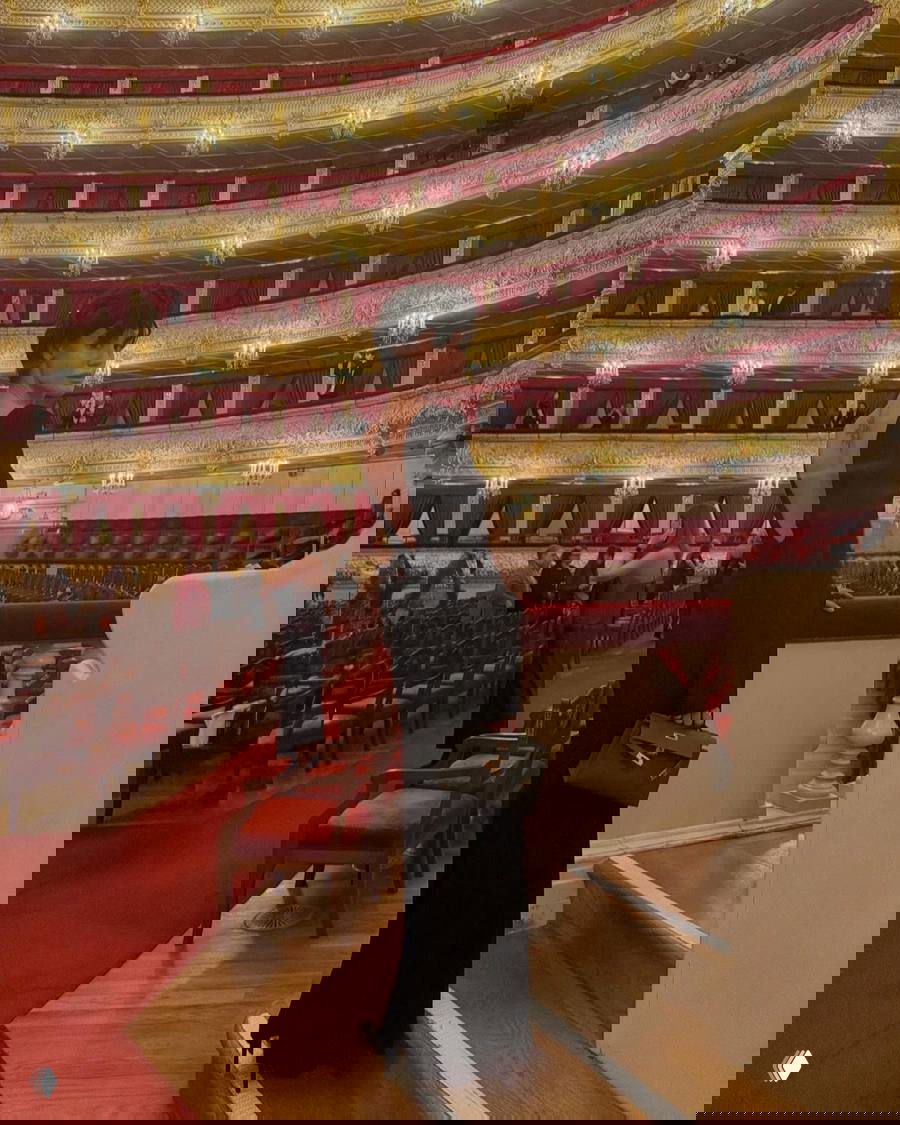 Широкий план интерьера Большого театра с гостями в вечерних нарядах и множеством лож, золотыми деталями и люстрами над залом.