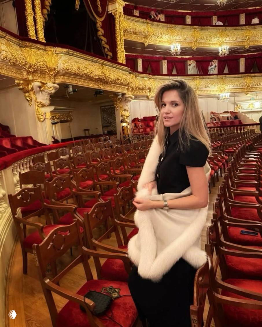 Женщина в платье с открытой спиной стоит у ступеней внутри Большого театра, позади виден ряд кресел и богато украшенный зал.