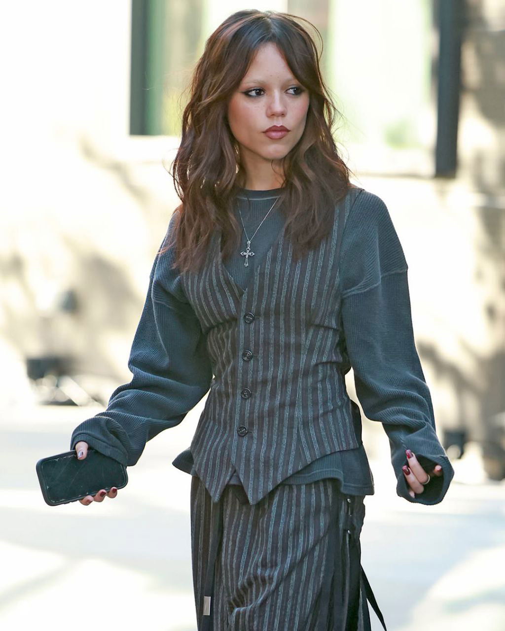 Крупный план Дженны Ортеги в повседневном образе: распущенные тёмные волосы, крест на цепочке и натуральный макияж на светлом фоне.