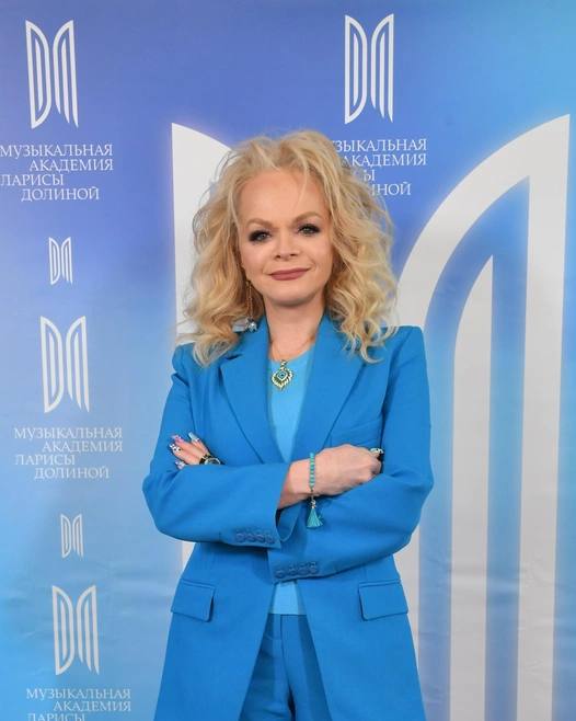 Женщина в голубом костюме позирует на фоне баннера с логотипами; портрет по пояс, официальная обстановка.