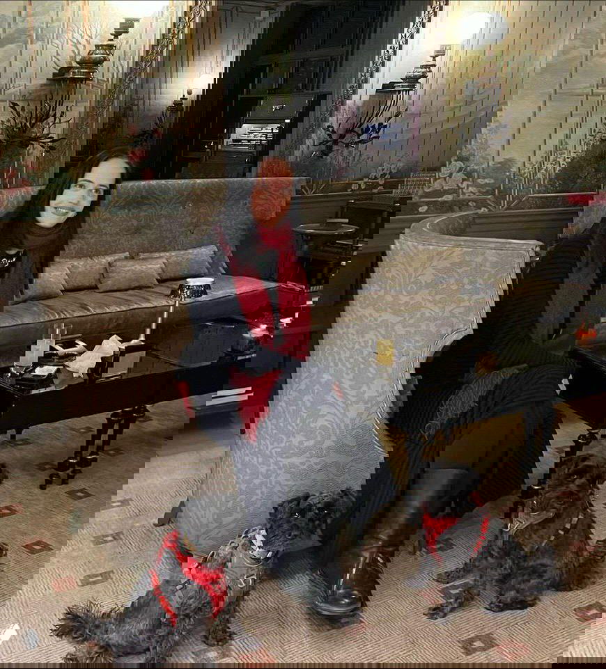 Ева Грин сидит в уютном холле рядом с двумя маленькими чёрными собаками в красных шлейках; старинный интерьер с диваном, столиком и декоративными панелями.