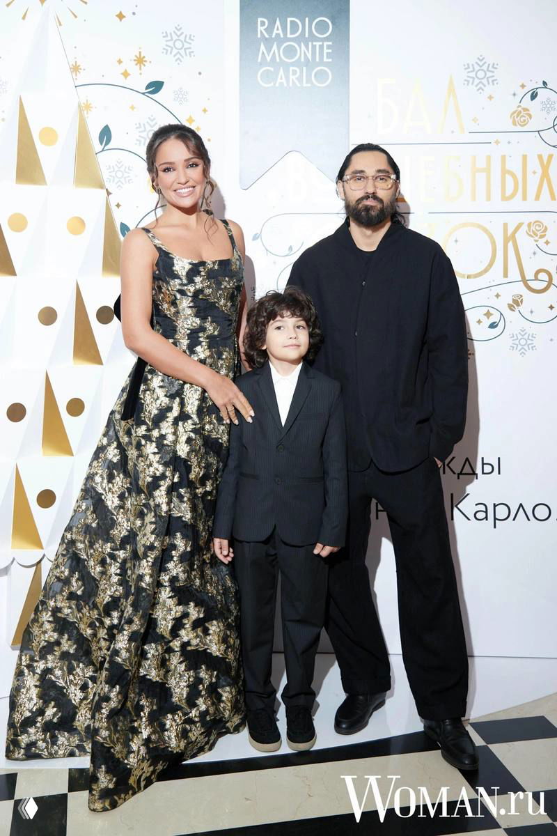 Семейный снимок: взрослые и ребёнок позируют в вечерних нарядах на фоне фирменного оформления бала «Волшебных сказок» Radio Monte Carlo.