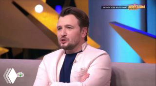 Крупный план телеведущего в студии шоу: мужчина сидит на диване в светлой пиджаке, фон с декорациями и логотипом передачи.