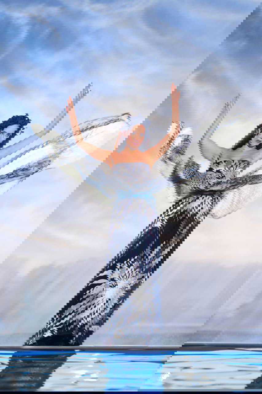 Женщина в длинном платье с развевающейся тканью, позирующая над водой на фоне неба; образ в морской эстетике съемки календаря.