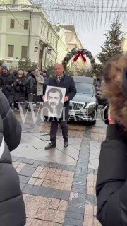 Мужчина держит портрет Игоря Золотовицкого на прощальной церемонии у МХТ, вокруг люди, катафалк и зимняя городская сцена, водяной знак Woman.ru