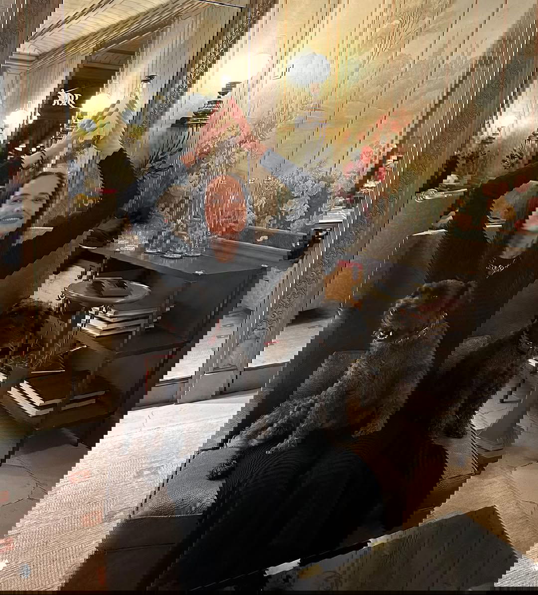 Ева Грин улыбается с бутылкой над головой, двое чёрных собак сидят у неё на коленях в элегантном зале с зеркалами и настенными панелями.