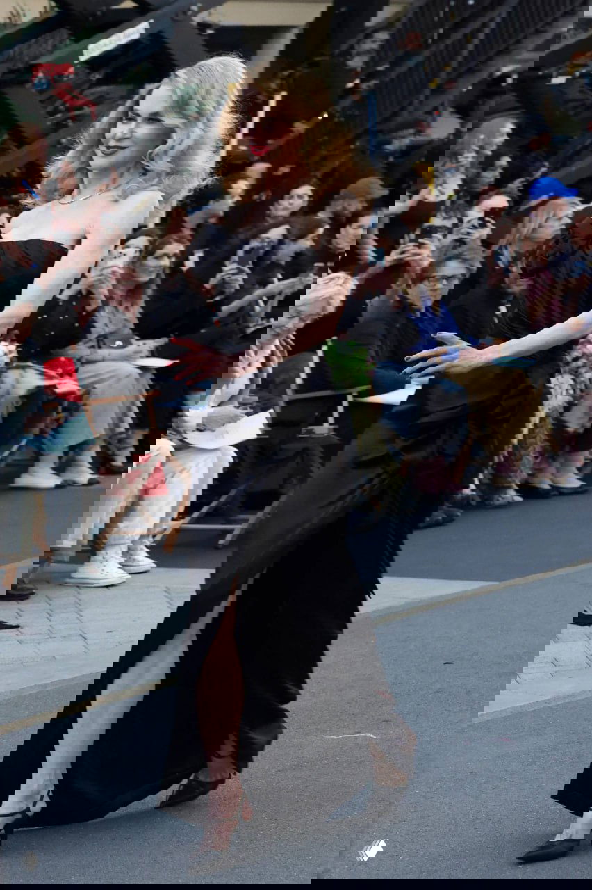 Женщина в чёрном платье идёт по подиуму модного показа, вокруг сидит публика и фотографы фиксируют выход.