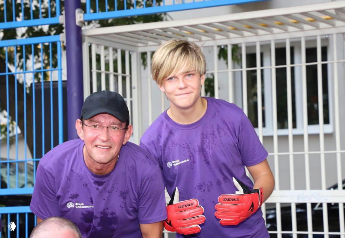 На фото мужчина и подросток в спортивной форме позируют после тренировки; они улыбаются на фоне ограждения и спортивного оборудования на улице.