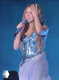 Крупный план Шакиры в сценическом образе с микрофоном и развивающимися волосами: момент выступления и эмоции певицы на концерте.