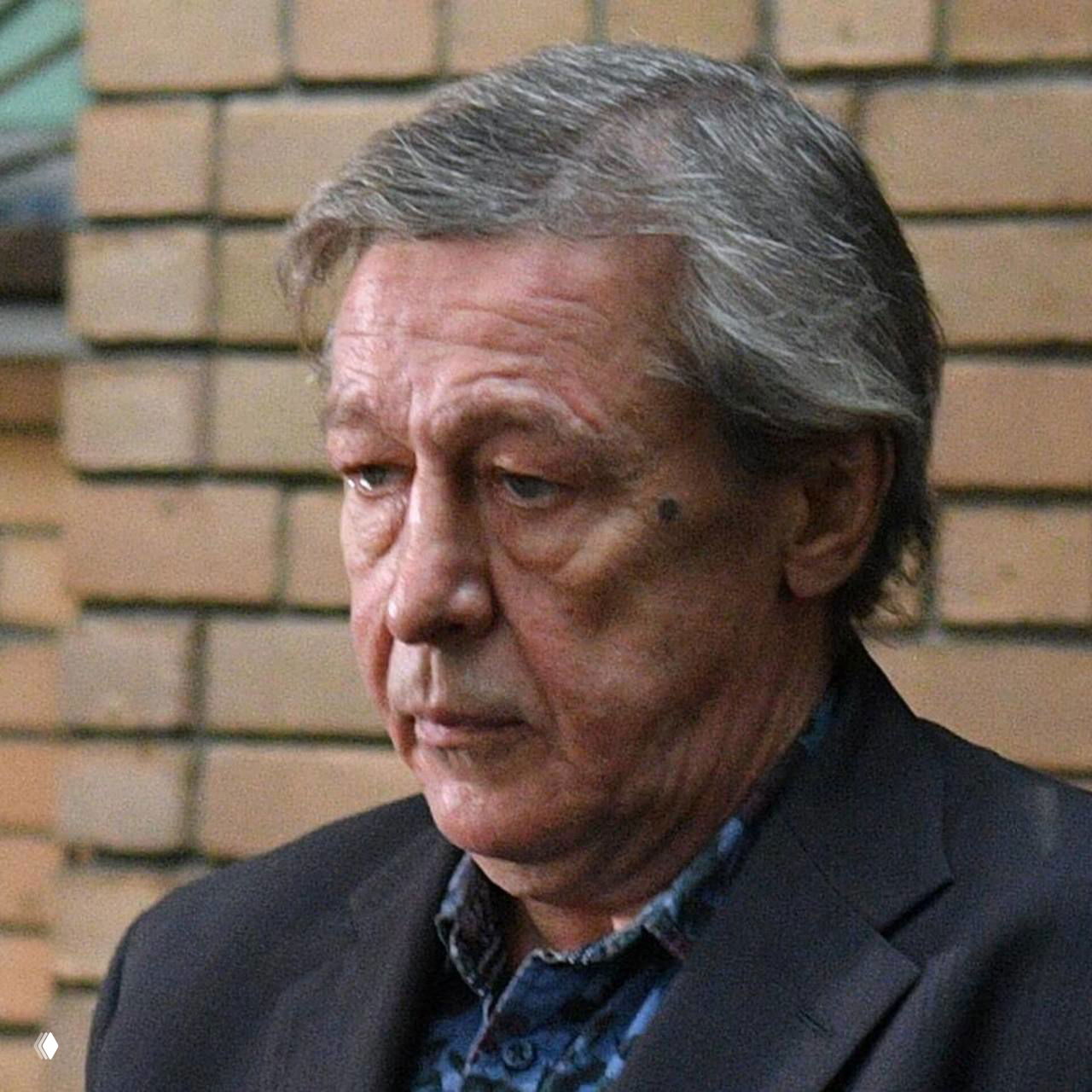 Крупный портрет пожилого мужчины в тёмном пиджаке на фоне кирпичной стены, крупный план лица и выражение серьёзности.