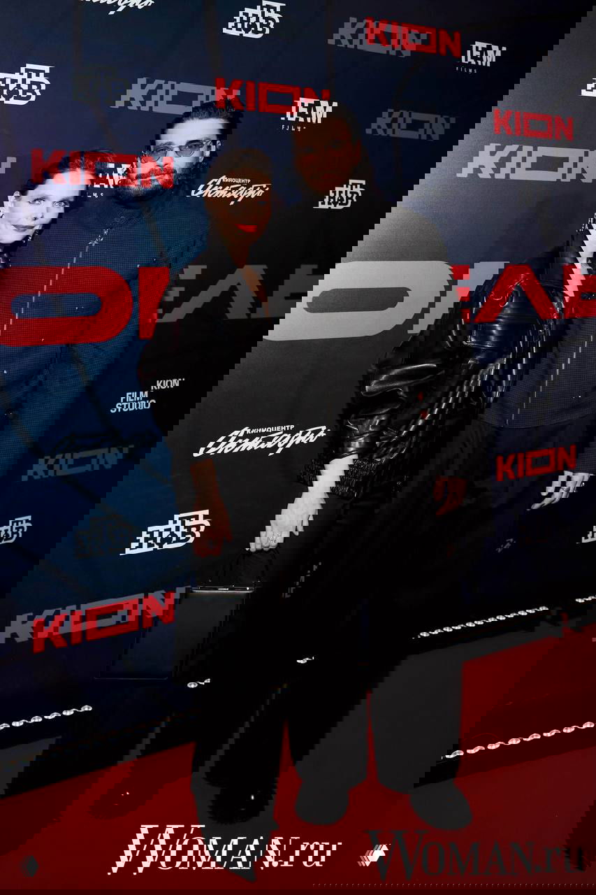 Фото пары на красной дорожке: женщина в чёрном наряде рядом с мужчиной, общий кадр у бренд‑волла кинопоказа с логотипами и спонсорами.