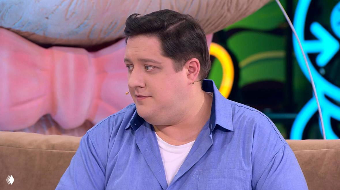 Крупный план мужчины в студийной телепрограмме в голубой рубашке; виден профиль и часть яркого студийного декора на заднем плане.