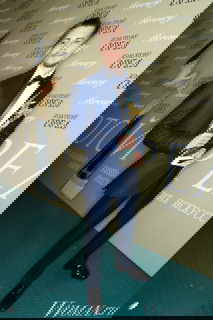 Иван Янковский в костюме на церемонии с наградой «Золотой орел», он держит статуэтку на фоне брендовой фотозоны мероприятия.