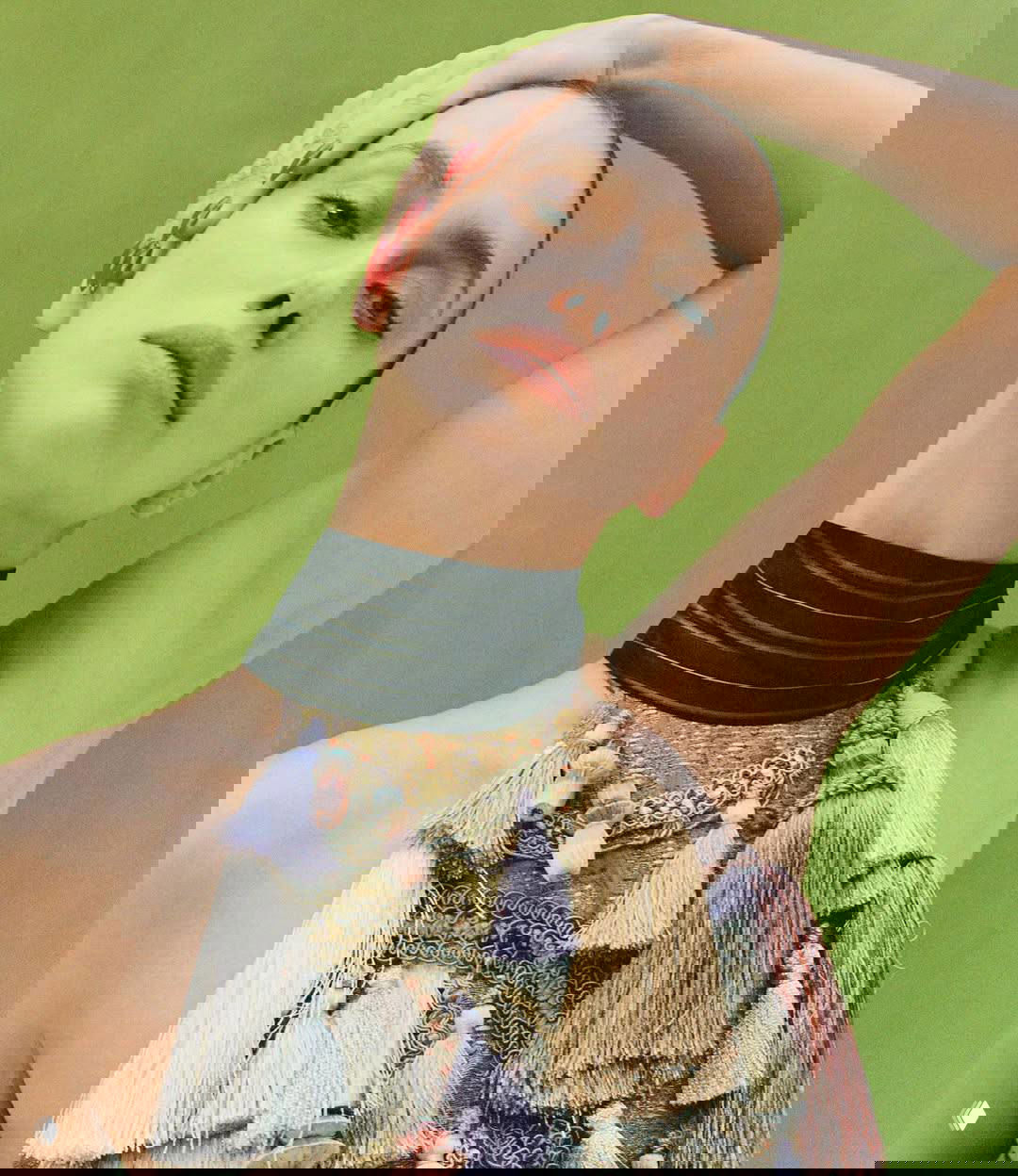 Крупный план модели на зелёном фоне с выразительными украшениями из кисточек и естественным макияжем, текстурная деталь одежды.