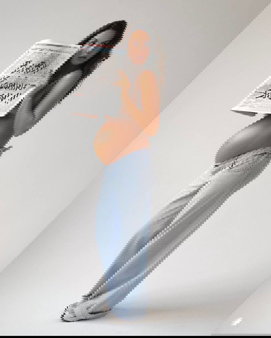 Стройная беременная модель в боковом профиле: джинсы на талии, обнажённый живот и газета с надписью о скором рождении.