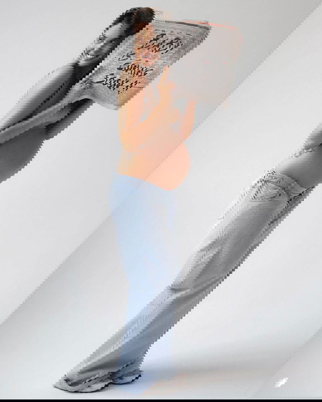 Беременная женщина улыбается и держит газету с надписью о скором появлении ребёнка, полный рост на нейтральном фоне.