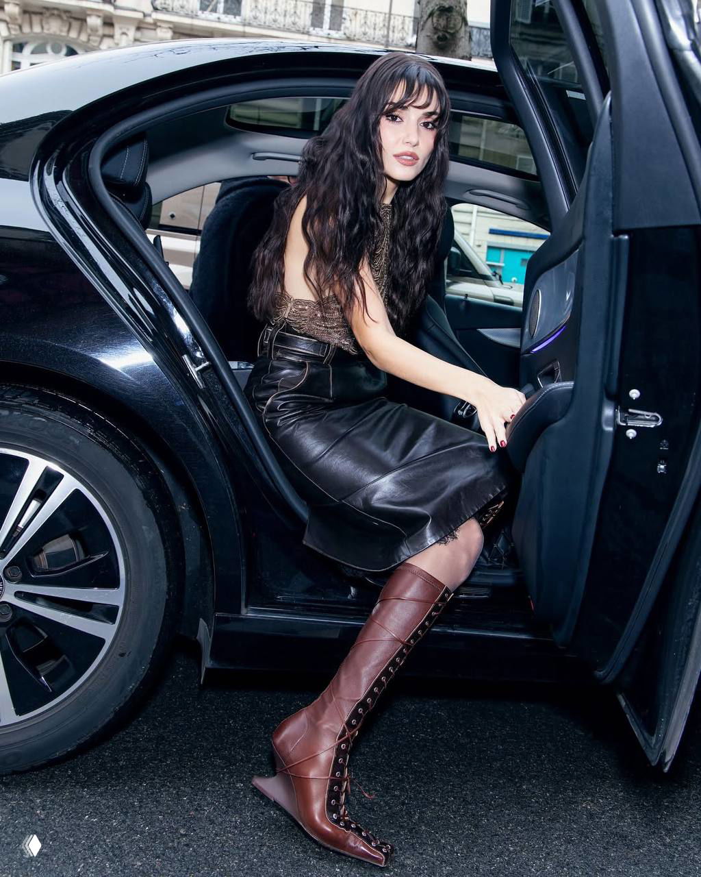 Женщина выходит из чёрного автомобиля в высоких коричневых ботфортах и кожаной юбке — кадр у curbside во время мероприятия.