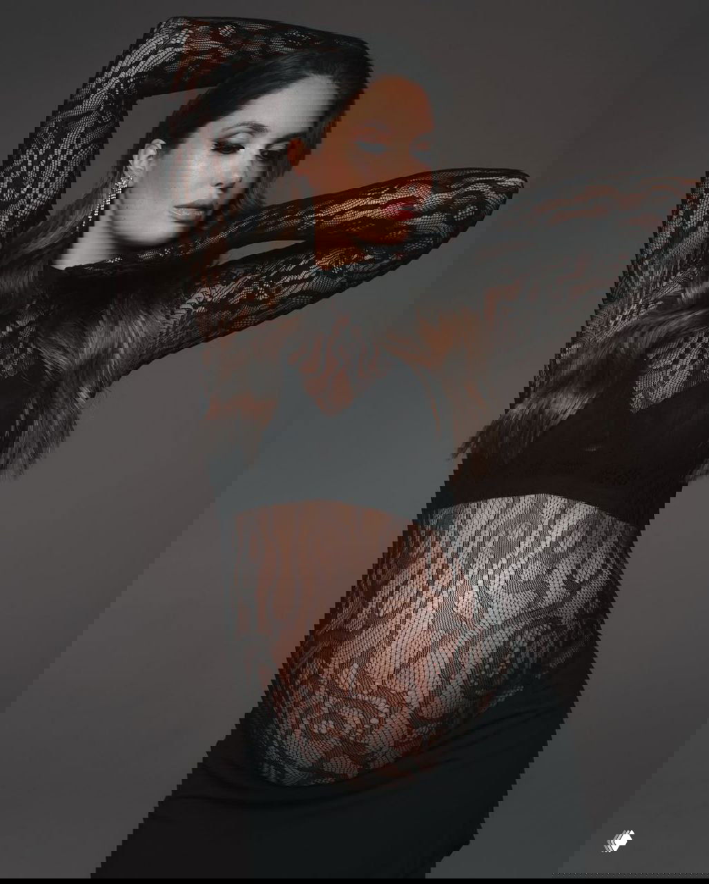 Агата Муцениеце в студийной фотосессии в черном кружевном платье, видно округлый беременный живот, поза элегантная и спокойная в портрете