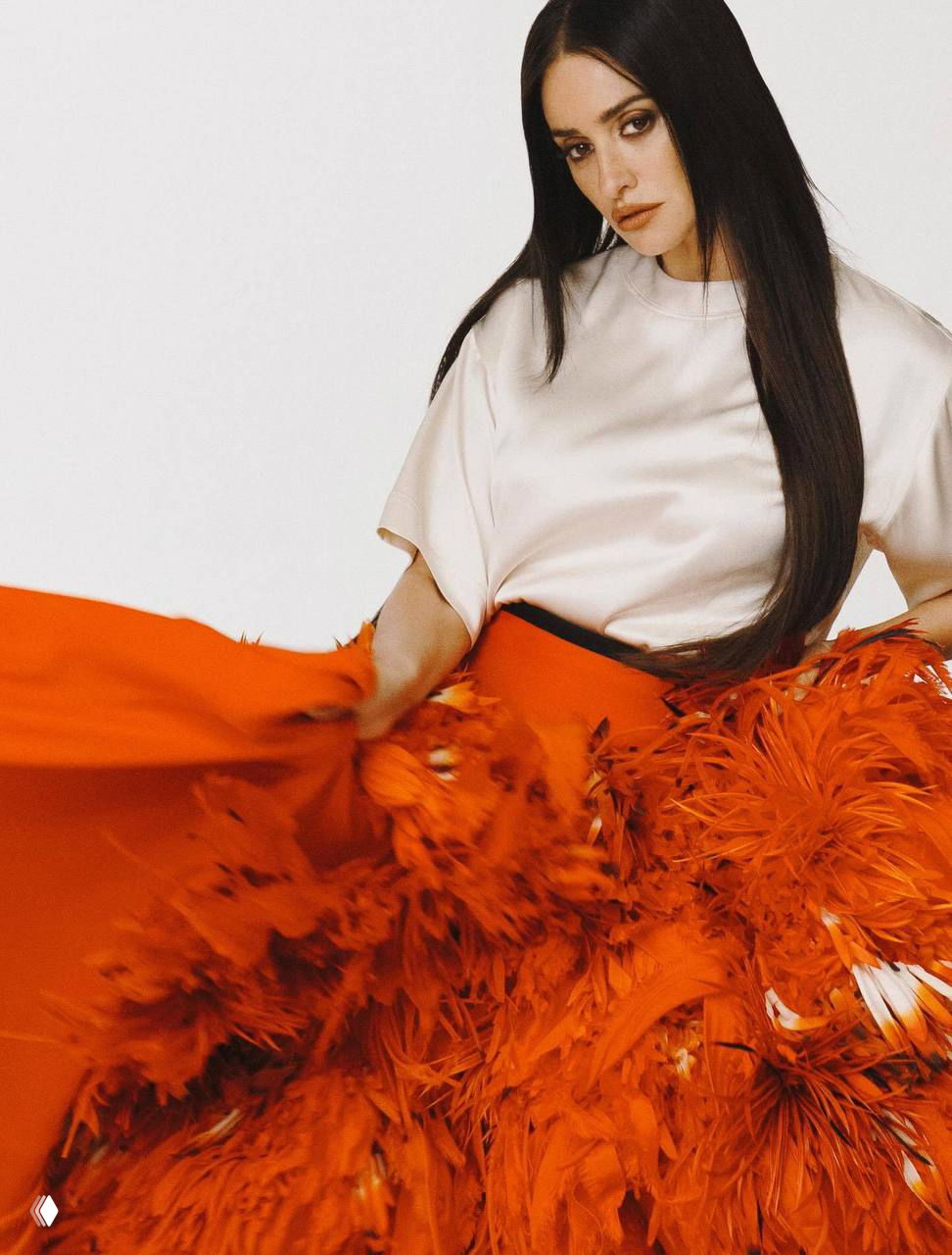 Динамичный кадр: женщина скользит рукой по пышной оранжевой юбке с перьями, текстура ткани подчёркивает движение.