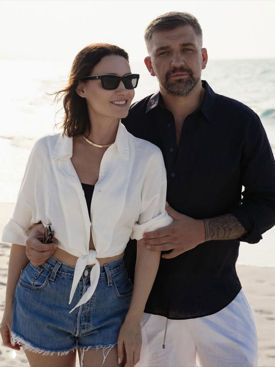 Пара стоит на пляже у кромки воды: мужчина в тёмной рубашке и женщина в белой рубашке с завязанным краем и джинсовыми шортами.