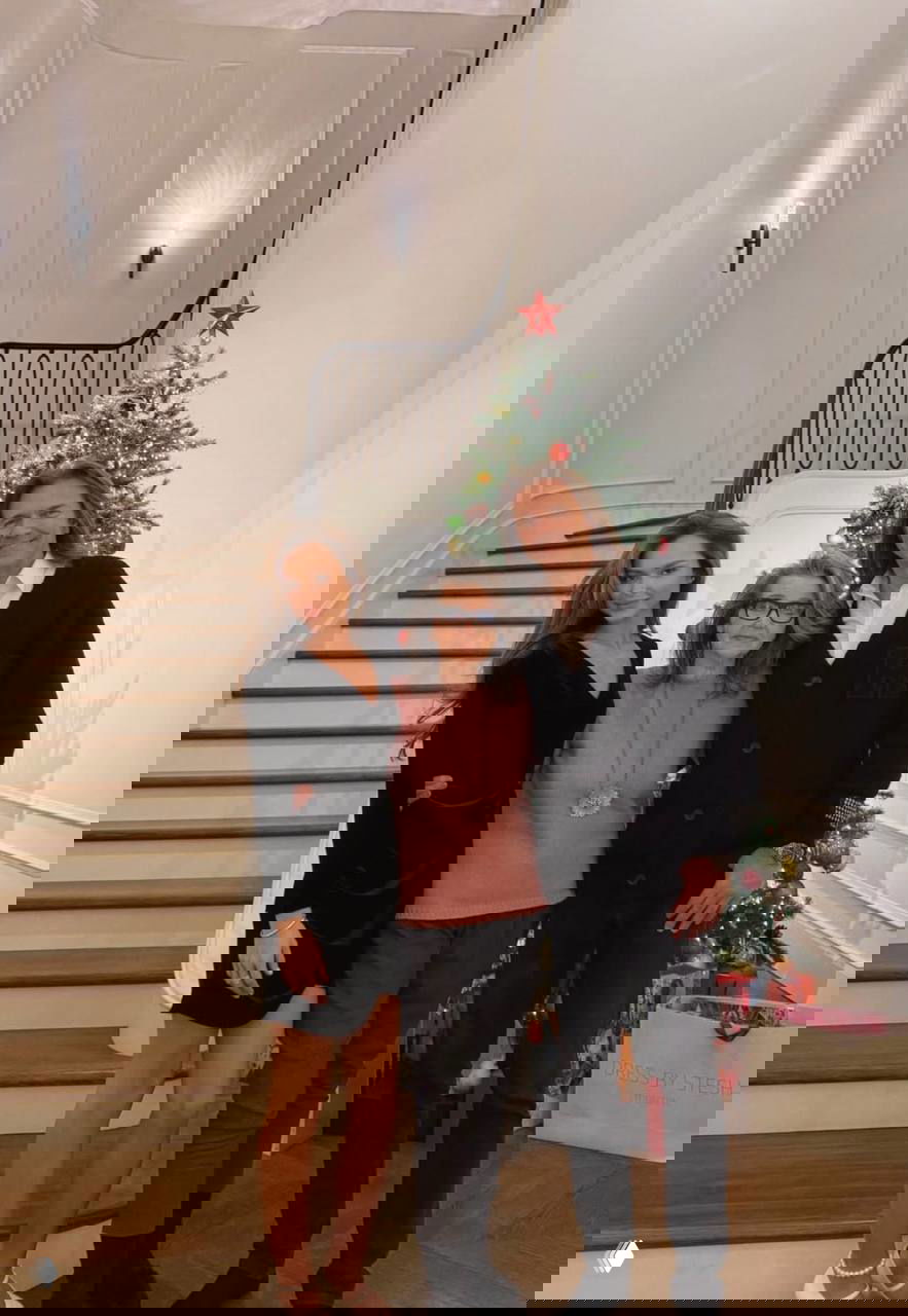 Три поколения на лестнице рядом с рождественской ёлкой: молодая женщина, пожилая родственница и мужчина позируют в доме с подарками.