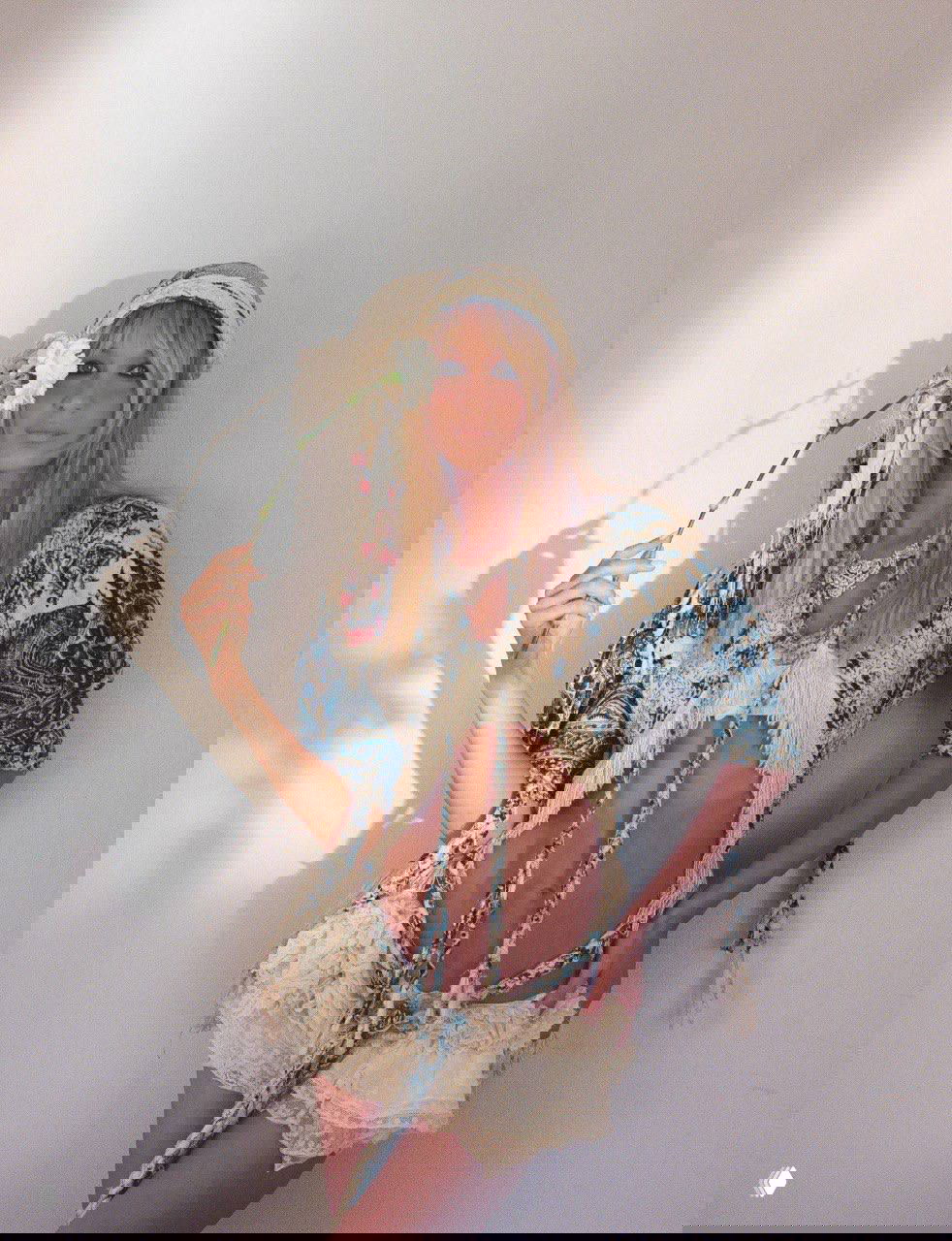 Белла Хадид в бохо-платье и головном уборе держит цветок, съёмка перед белой стеной с мягким естественным светом.