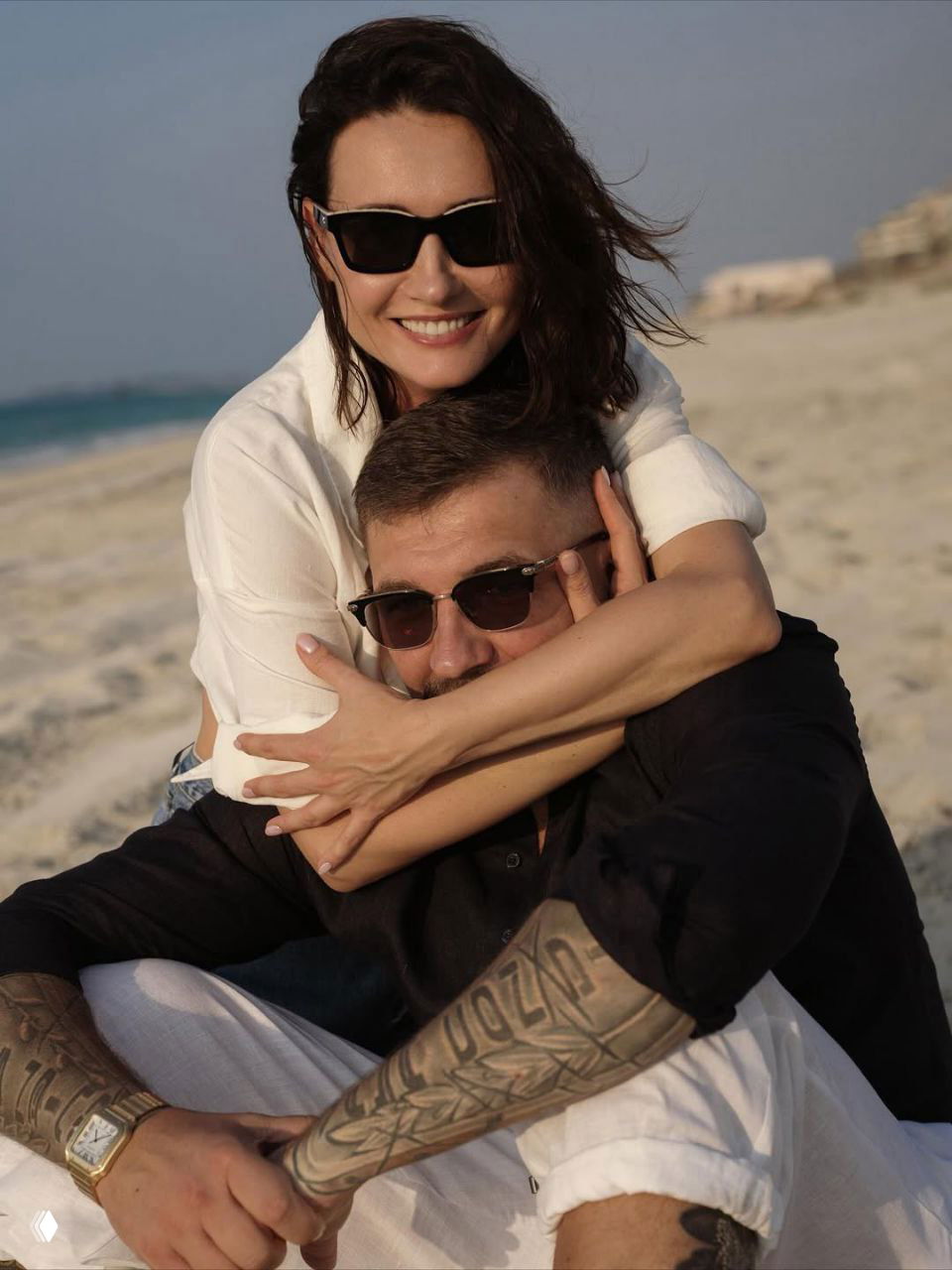 Женщина в солнечных очках обнимает мужчину с татуировками: они сидят на покрывале на песке, вокруг морской пейзаж и закатный свет.