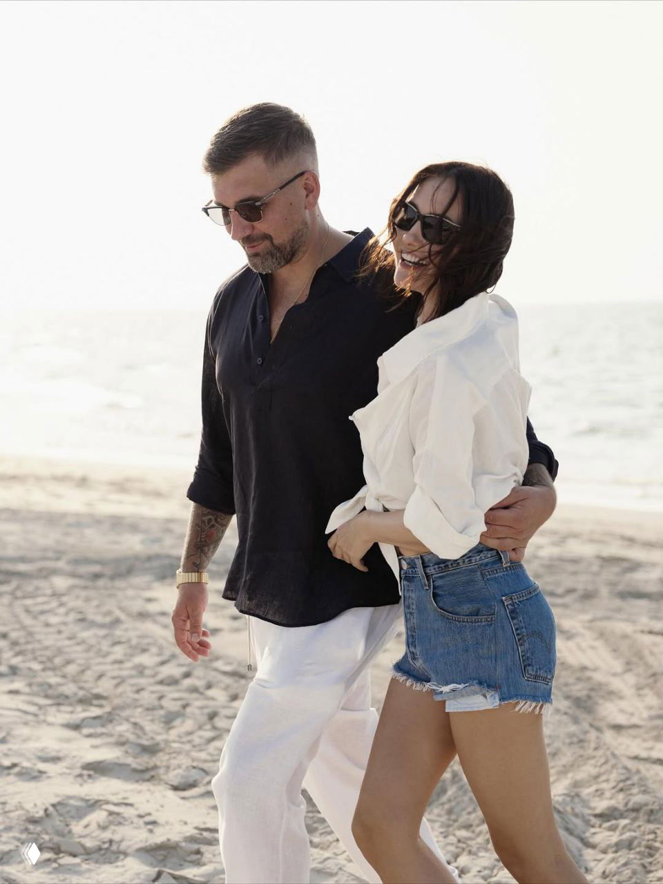 Кадр пары, проходящей по песку возле прибоя: она улыбается в солнечных очках, он держит её под руку, пляж и море вокруг.