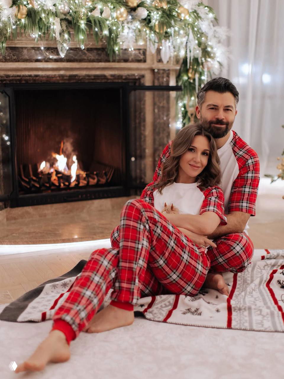 Пара в праздничной пижаме сидит у камина с горящим огнём и рождественским декором в комнате — уютная домашняя сцена.
