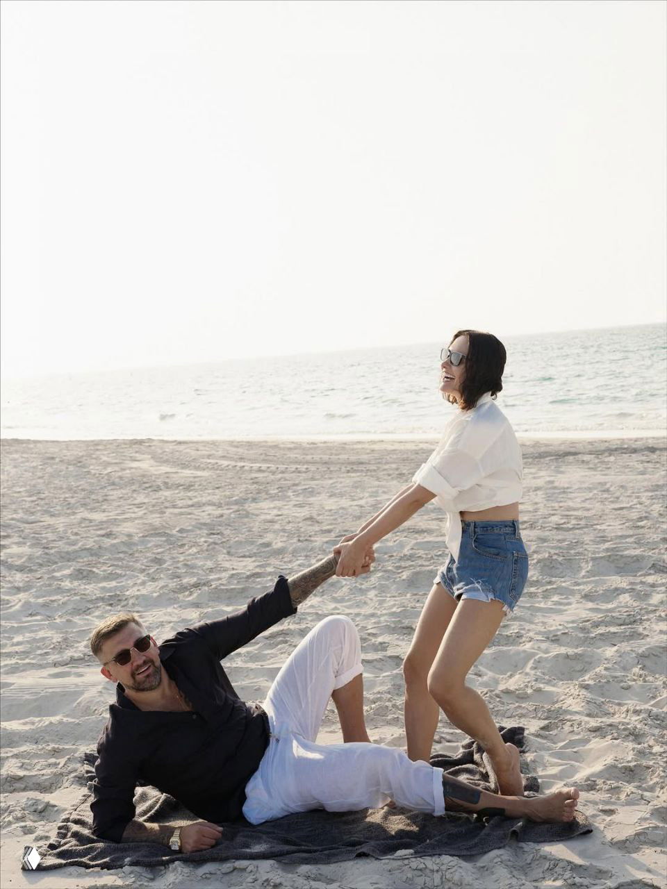 Мужчина и женщина играют у воды на песке: она тянет его за руку, они улыбаются, вокруг светлый морской фон и песчаный берег.