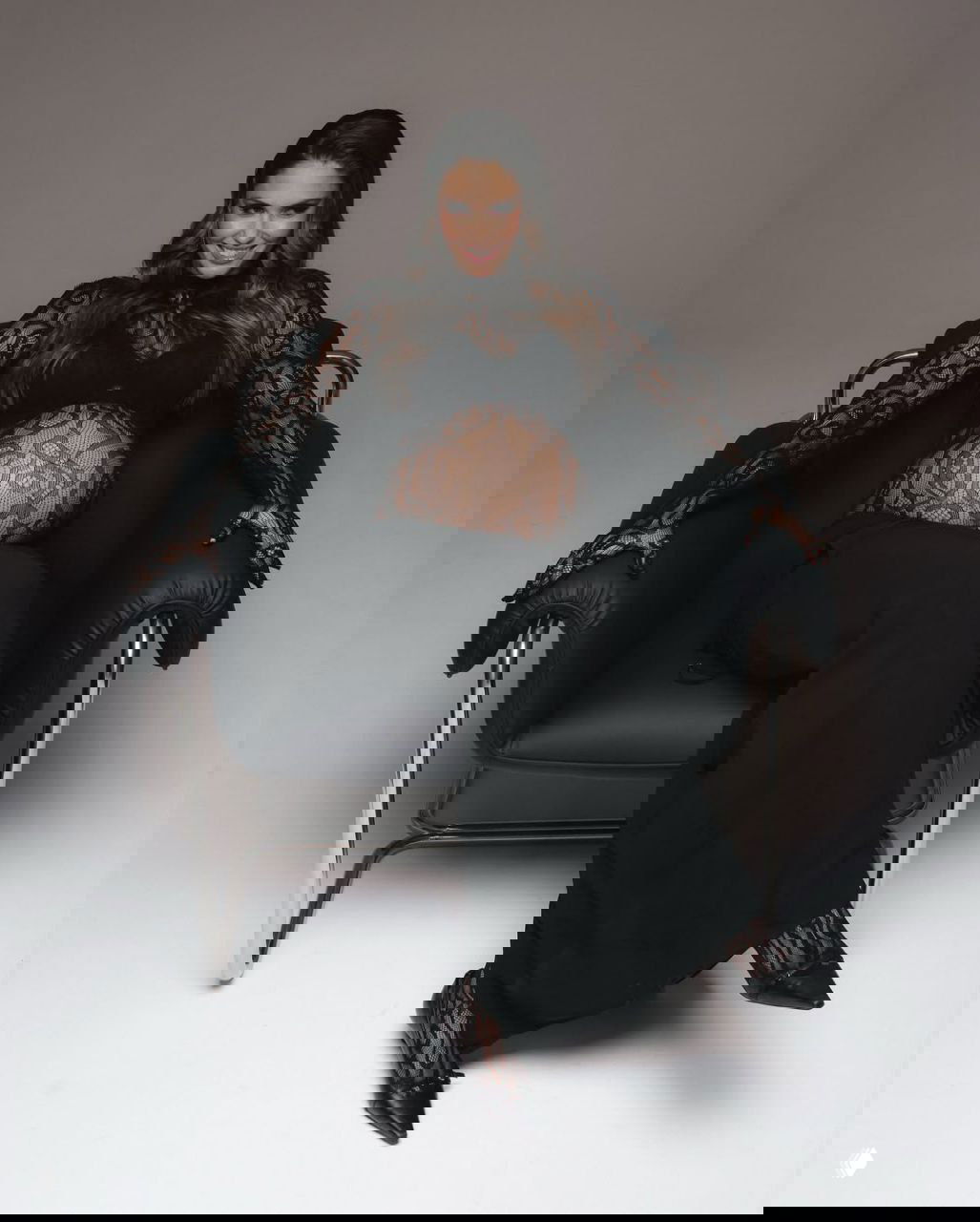 Агата Муцениеце сидит в кресле в кружевном наряде, расслабленная поза, улыбка и подчеркнутая форма живота в студийном свете