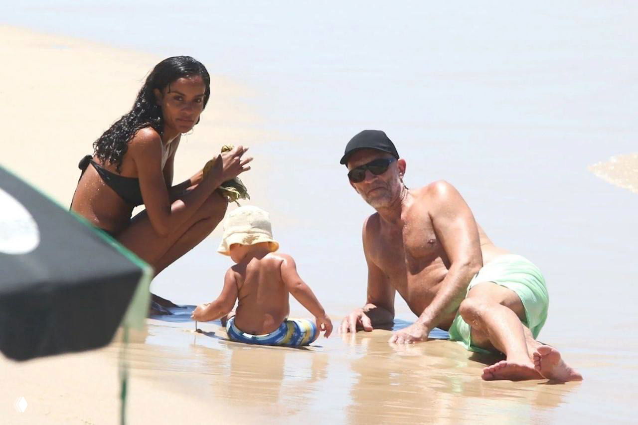 Женщина в чёрном бикини и мужчина с кепкой сидят у кромки воды рядом с маленьким ребёнком в панаме, играющим в песке у прибоя.