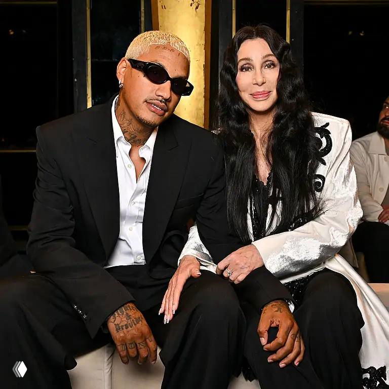 Пожилая женщина и молодой мужчина сидят рядом в торжественной обстановке, оба в вечерней одежде, улыбаются и позируют для фото.