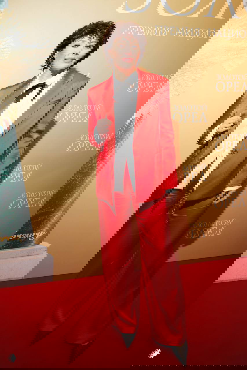 Екатерина Волкова в ярком красном костюме с белой блузой и галстуком позирует на фоне золотого баннера церемонии «Золотой орёл».