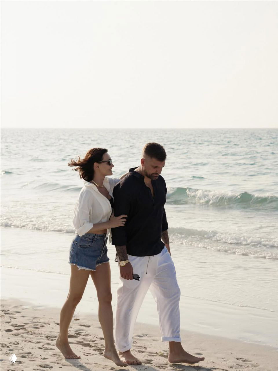 Мужчина и женщина идут по пляжу, держась за руки; на заднем плане волны, они в расслабленных летних образах, сцена динамичная.
