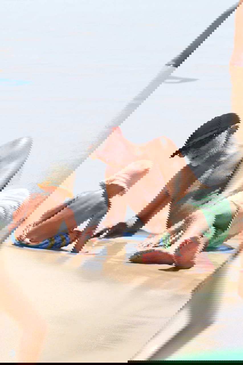 Мужчина в кепке лежит у берега рядом с малышом, который сидит и играет с мокрым песком; солнечный пляж и лёгкая волна.