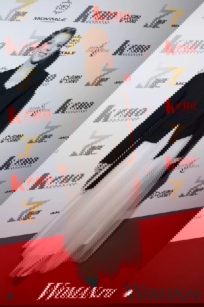 Женщина в вечернем образе на красной дорожке у баннера «Кино Репортер», полный рост, светлая многослойная юбка и тёмный верх наряда.